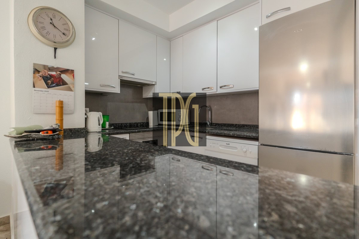 Modern white kitchen with glossy cabinets, dark granite counters, a stainless steel fridge and dishwasher, a wall clock and calendar on the wall, and a kettle on the counter.