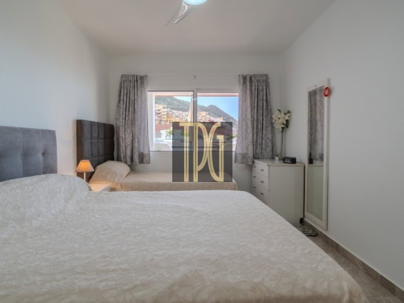 Hotel bedroom with two twin beds and grey tufted headboards; window view of buildings and hills, dresser with flowers and a full-length mirror nearby.