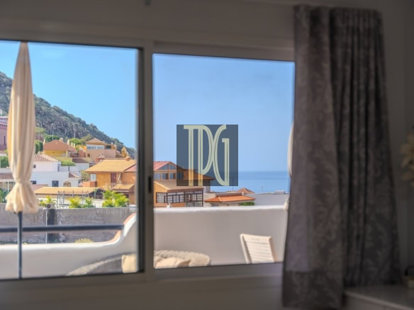 View from a living room window showing a balcony with a beige umbrella, pastel hillside houses, and the blue sea beyond.