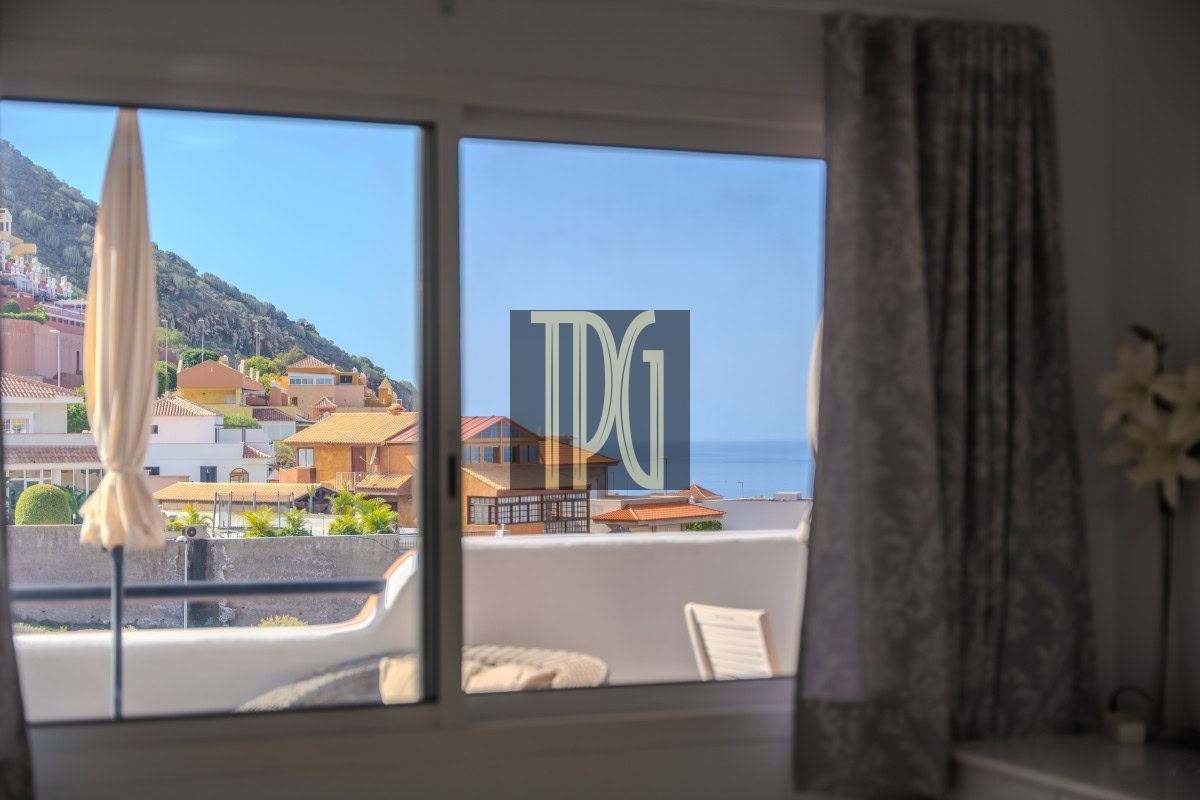 View from a living room window showing a balcony with a beige umbrella, pastel hillside houses, and the blue sea beyond.