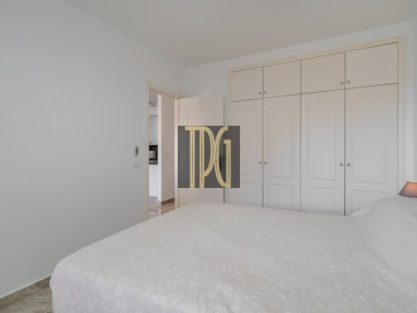 Bright bedroom with a large white paneled wardrobe along the wall and a white bed in the foreground, doorway to a hallway on the left.