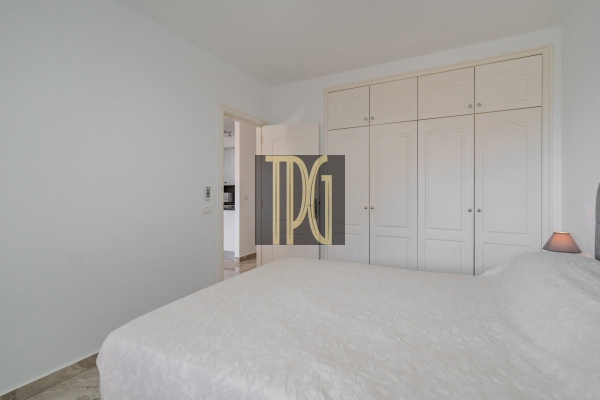 Bright bedroom with a large white paneled wardrobe along the wall and a white bed in the foreground, doorway to a hallway on the left.
