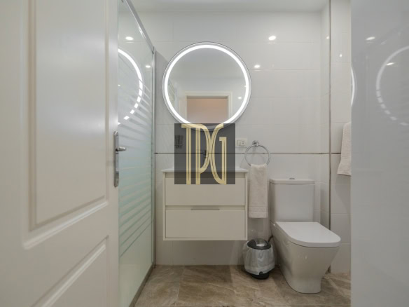 Modern white bathroom with a circular backlit mirror above a floating vanity, glass shower door, towel ring, and toilet
