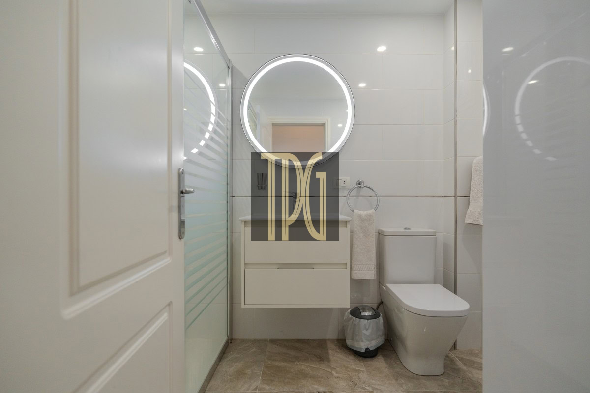 Modern white bathroom with a circular backlit mirror above a floating vanity, glass shower door, towel ring, and toilet