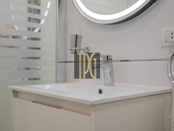 White bathroom vanity with a rectangular sink and chrome faucet, round lighted mirror above, frosted glass shower screen on the left, towel ring and outlet on the right.