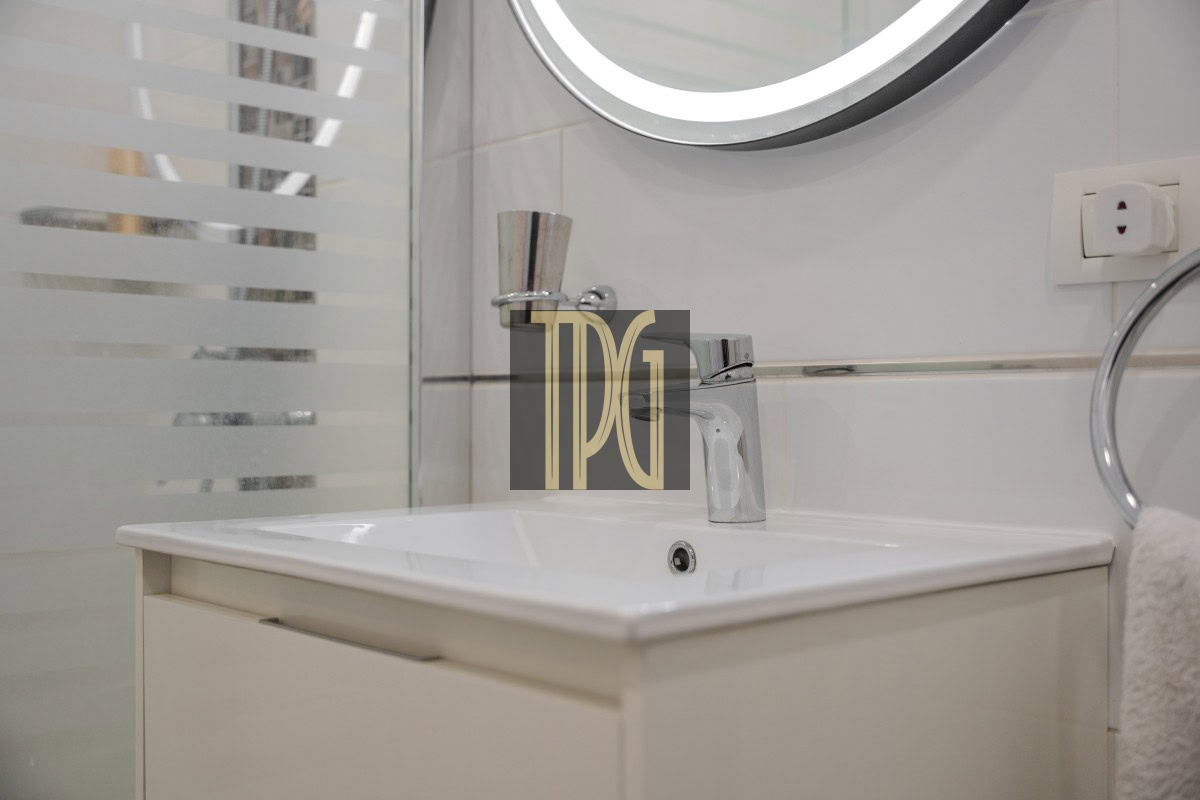 White bathroom vanity with a rectangular sink and chrome faucet, round lighted mirror above, frosted glass shower screen on the left, towel ring and outlet on the right.