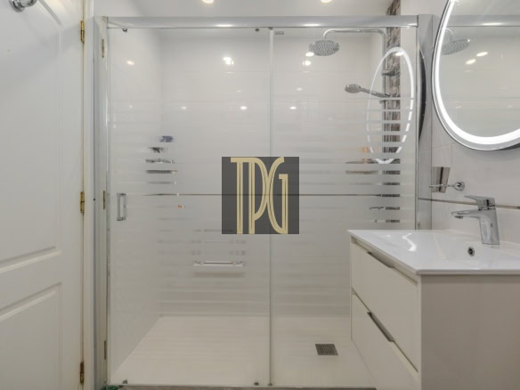 Modern white bathroom featuring a glass shower enclosure with frosted stripes and a white vanity with a round backlit mirror; TDG watermark centered on the glass door.