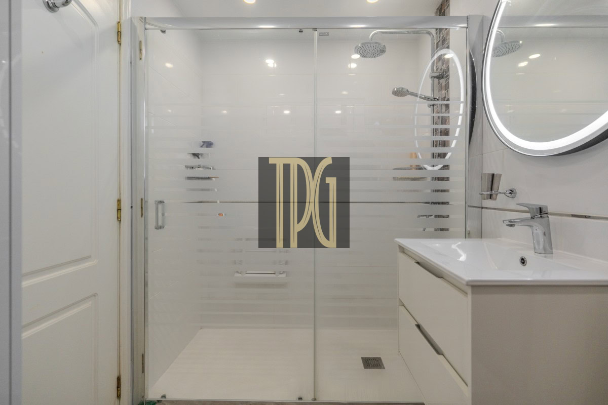 Modern white bathroom featuring a glass shower enclosure with frosted stripes and a white vanity with a round backlit mirror; TDG watermark centered on the glass door.