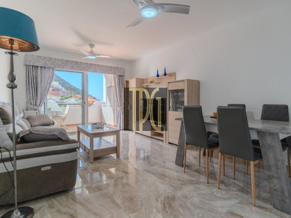 Bright open-plan living and dining room with a beige sofa, marble dining table, and balcony doors showing a town and hills outside.