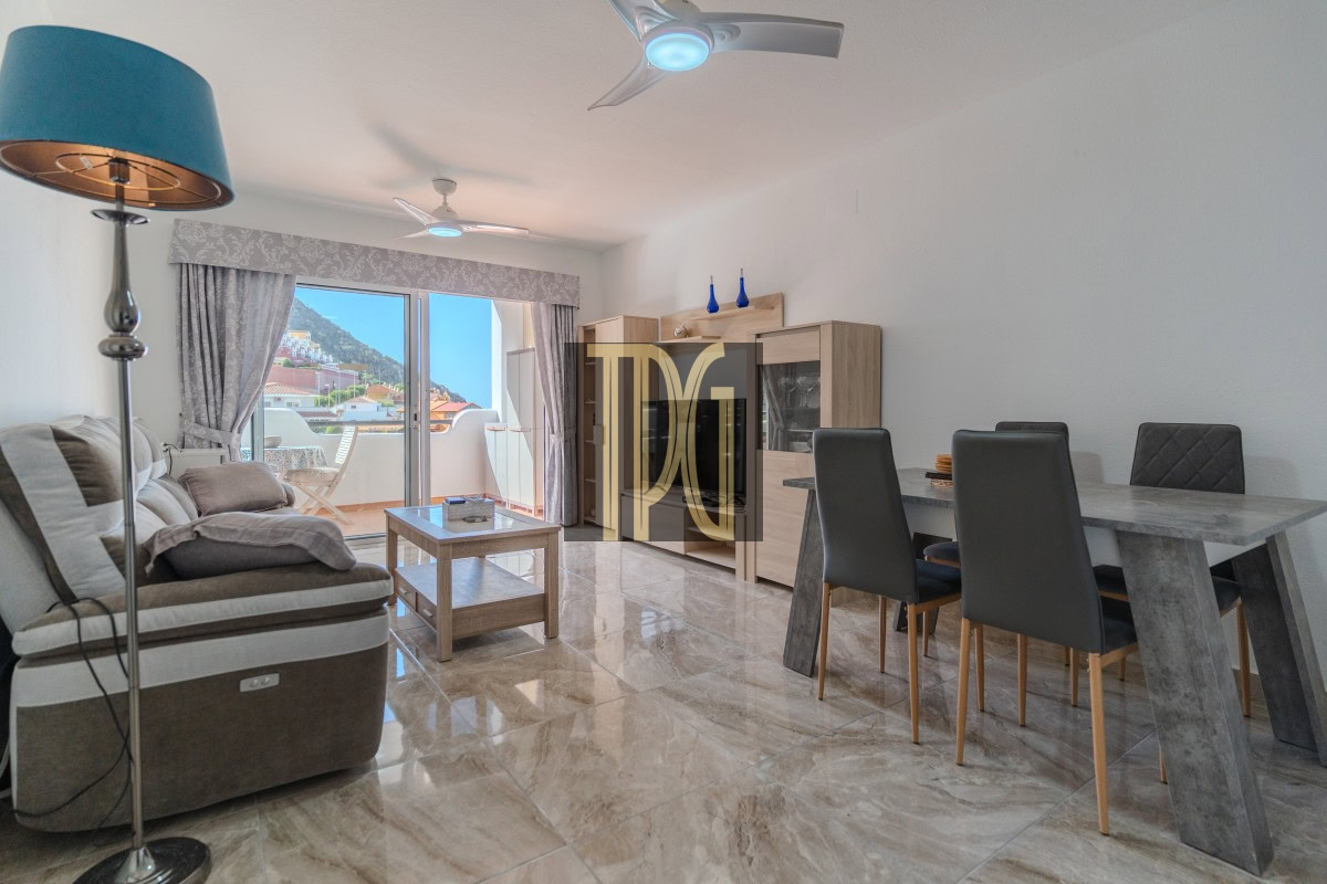 Bright open-plan living and dining room with a beige sofa, marble dining table, and balcony doors showing a town and hills outside.