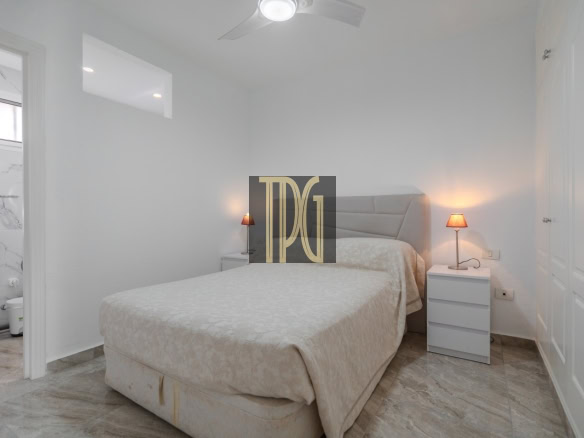 Bright white bedroom with a queen bed, two nightstands, and a ceiling fan; bathroom entry visible on the left.