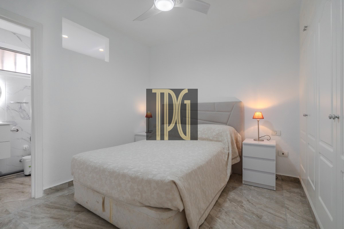 Bright white bedroom with a queen bed, two nightstands, and a ceiling fan; bathroom entry visible on the left.