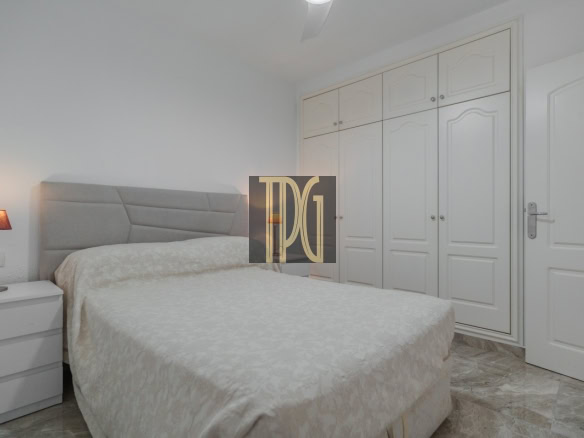 Bedroom with a large bed, beige quilt, padded headboard, white nightstand, and a red bedside lamp.
