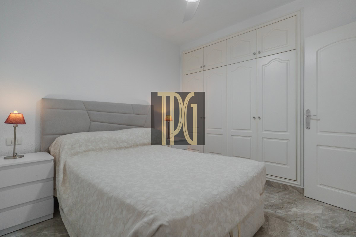 Bedroom with a large bed, beige quilt, padded headboard, white nightstand, and a red bedside lamp.