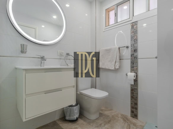 Bright modern bathroom with a white vanity, round illuminated mirror, and wall-mounted faucet opposite a toilet; towel rack and mosaic tile accent along the wall; small window above.
