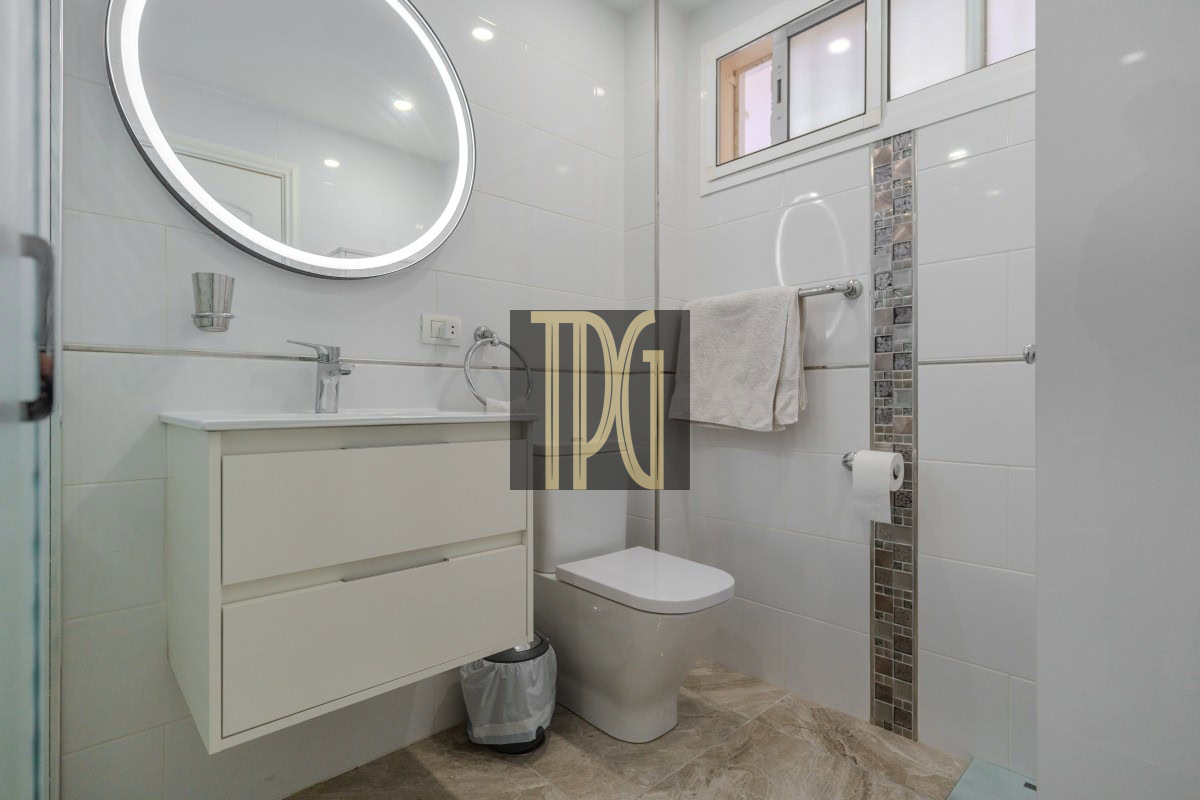 Bright modern bathroom with a white vanity, round illuminated mirror, and wall-mounted faucet opposite a toilet; towel rack and mosaic tile accent along the wall; small window above.