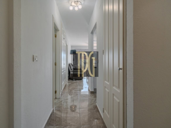 Bright hallway in a home with white walls and marble floor, opening to a living area with a sofa in the distance.