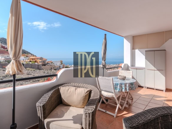 Sunlit balcony with wicker chair and beige cushion, a round table with a blue-patterned cloth, and an umbrella, overlooking a coastal town and the sea.