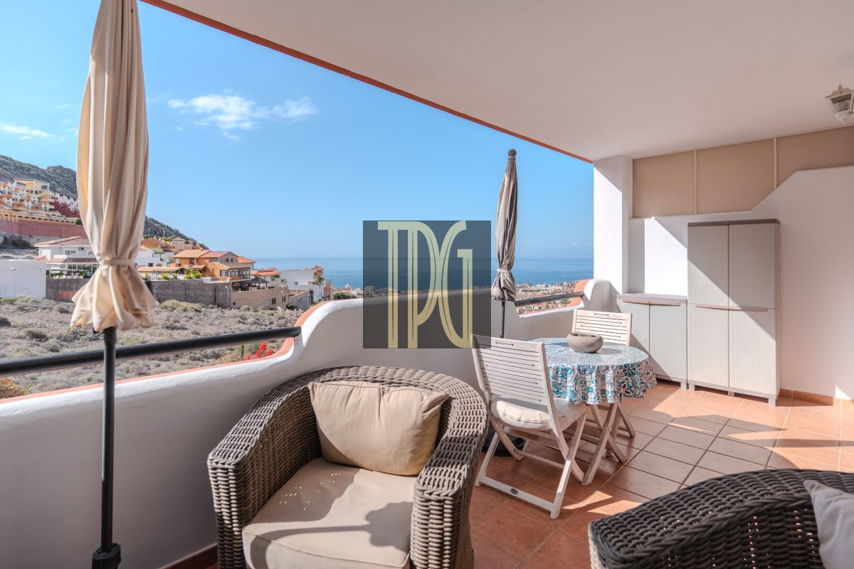 Sunlit balcony with wicker chair and beige cushion, a round table with a blue-patterned cloth, and an umbrella, overlooking a coastal town and the sea.