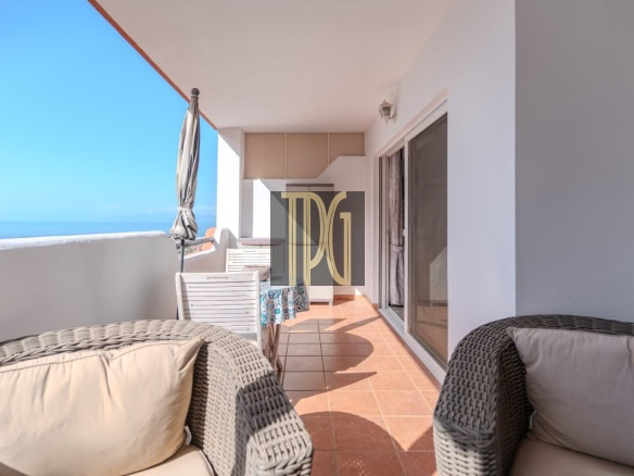 Sunlit ocean-side balcony with wicker chairs, cream cushions, and a glass door opening to an interior space; blue sea on the horizon and a closed umbrella. or A spacious terrace with terracotta tiles, woven rattan seating, and a view of the sea under a clear blue sky; sliding glass doors on the right.