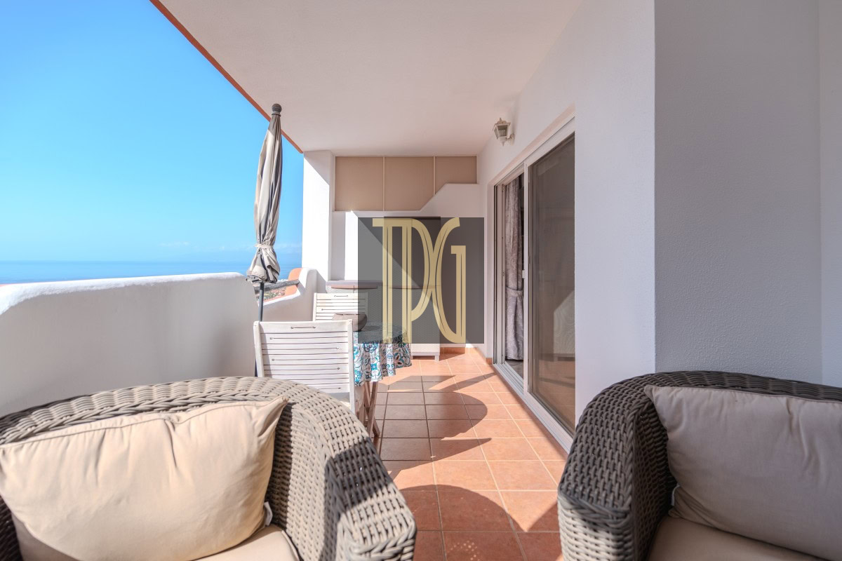 Sunlit ocean-side balcony with wicker chairs, cream cushions, and a glass door opening to an interior space; blue sea on the horizon and a closed umbrella. or A spacious terrace with terracotta tiles, woven rattan seating, and a view of the sea under a clear blue sky; sliding glass doors on the right.