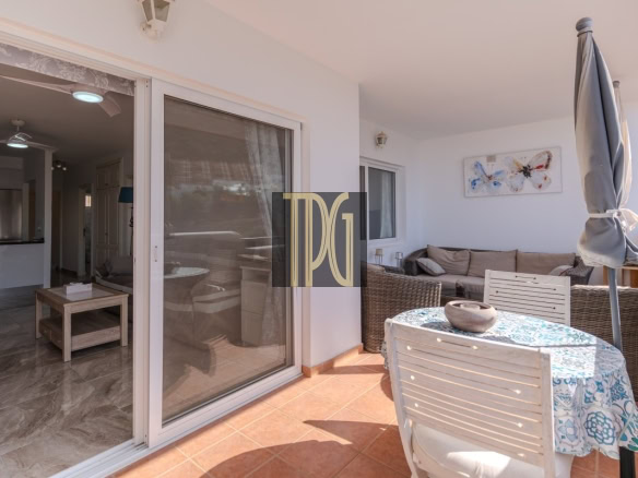 Outdoor patio with a round table covered by a blue-patterned tablecloth, white chairs, and a tall closed umbrella beside a wicker sofa. The scene is sunny and inviting.