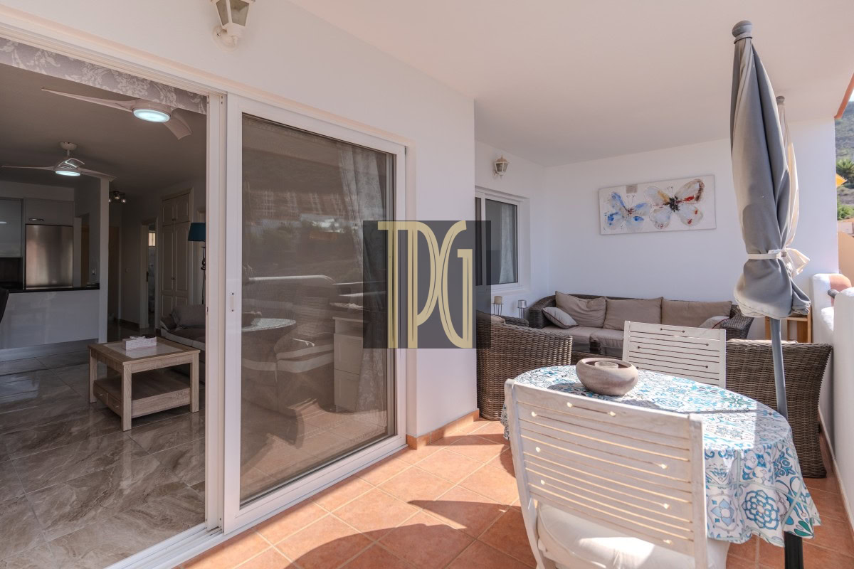 Outdoor patio with a round table covered by a blue-patterned tablecloth, white chairs, and a tall closed umbrella beside a wicker sofa. The scene is sunny and inviting.