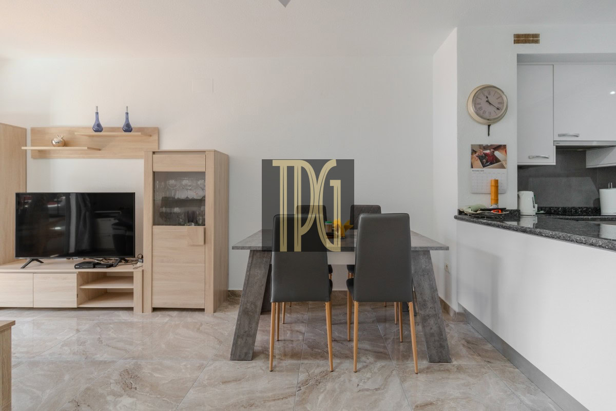 Open-plan living and dining area with a wooden TV cabinet, mounted TV, concrete-look dining table, and four gray chairs around it.