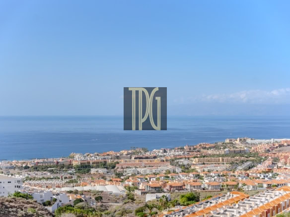 Coastal town with orange-roofed houses along the shore and calm sea under a clear blue sky, with a centered dark watermark logo reading TDG in the middle.