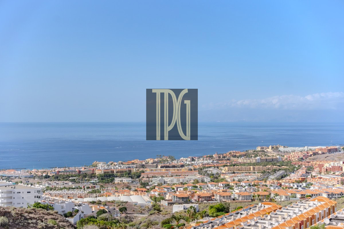 Coastal town with orange-roofed houses along the shore and calm sea under a clear blue sky, with a centered dark watermark logo reading TDG in the middle.