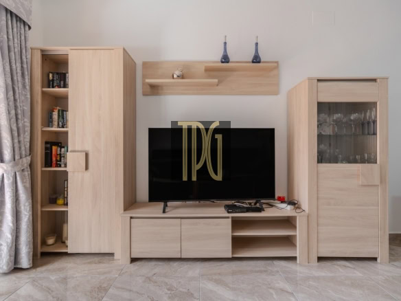 Light wood entertainment unit surrounding a flat-screen TV, with a tall bookcase on the left and a glass-front cabinet on the right, and curtains to the left.
