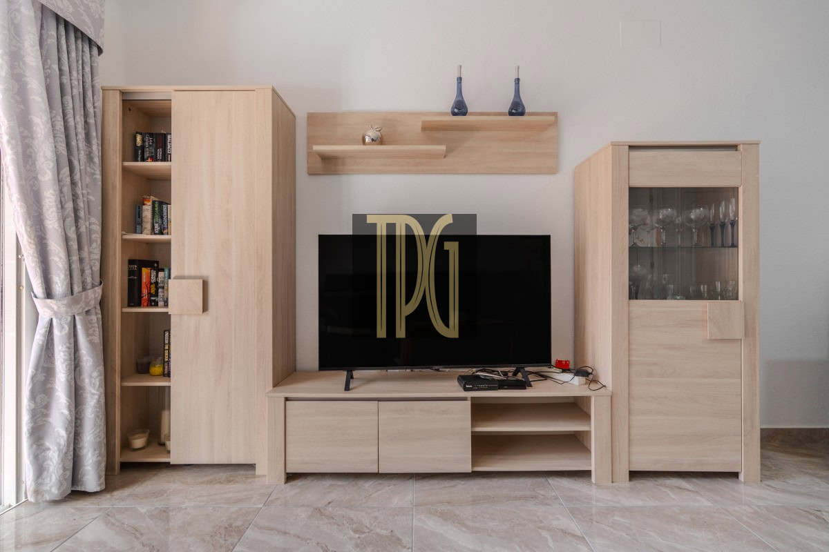 Light wood entertainment unit surrounding a flat-screen TV, with a tall bookcase on the left and a glass-front cabinet on the right, and curtains to the left.