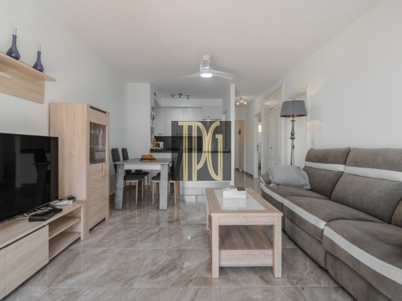 Open-plan living room with gray sectional sofa, coffee table, and TV stand; dining table and chairs visible toward the kitchen; beige tiled floor and neutral decor.