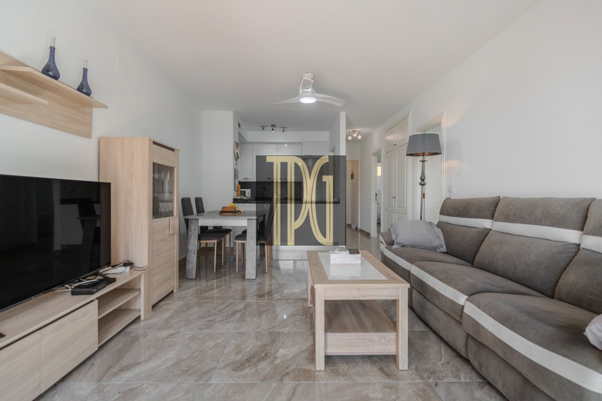 Open-plan living room with gray sectional sofa, coffee table, and TV stand; dining table and chairs visible toward the kitchen; beige tiled floor and neutral decor.