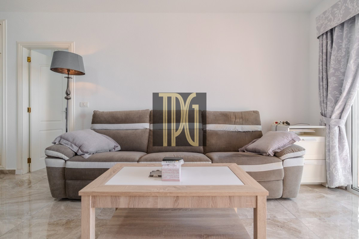 Bright living room with a beige sectional sofa, light wood coffee table, floor lamp on the left, and patterned curtains on the right.