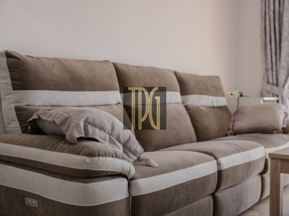 Brown upholstered three-seat sofa with light beige piping and a folded blanket, set in a bright living room with curtains at the window.