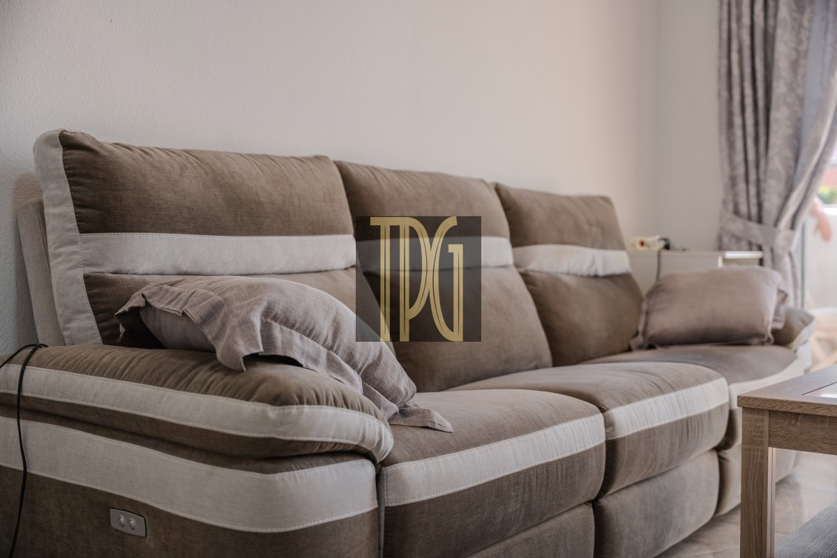 Brown upholstered three-seat sofa with light beige piping and a folded blanket, set in a bright living room with curtains at the window.