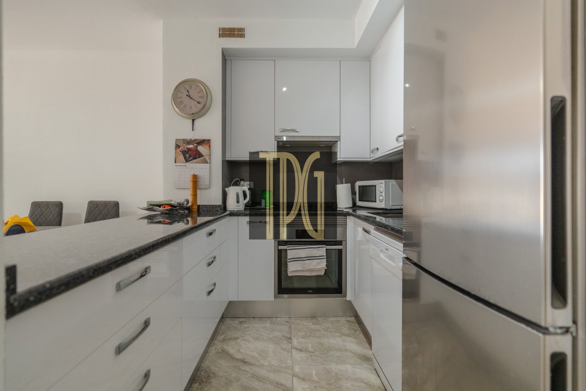 Modern white kitchen with granite countertops, built-in oven, microwave, and stainless fridge; wall clock and calendar above the counter.