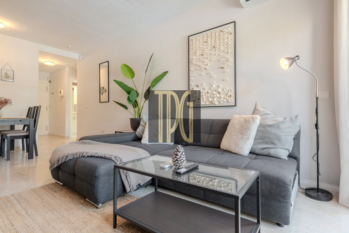 Modern living room with a dark gray sectional sofa, glass coffee table, and a tall floor lamp by the wall art. A potted plant and beige throw pillows complete the space.