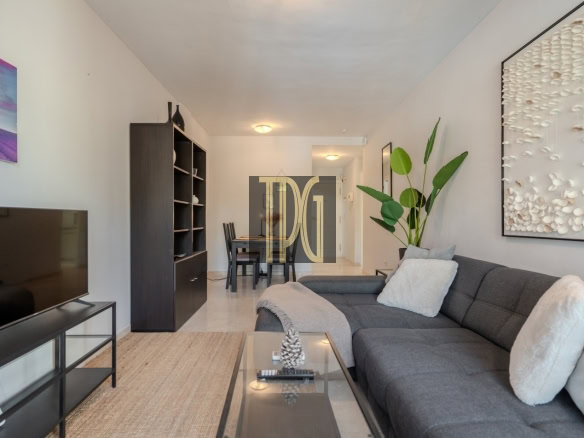 Modern living room with a gray sectional, glass coffee table, TV stand, and large wall art in a bright, open space with a plant nearby.