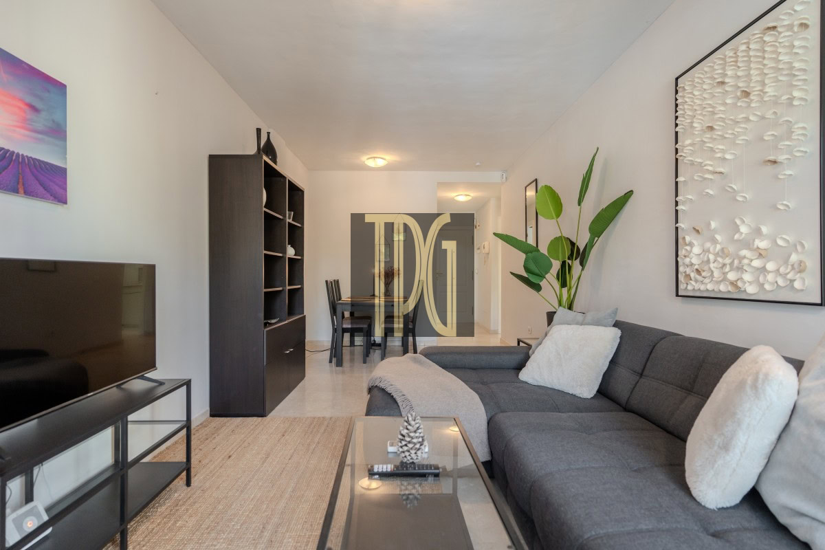 Modern living room with a gray sectional, glass coffee table, TV stand, and large wall art in a bright, open space with a plant nearby.