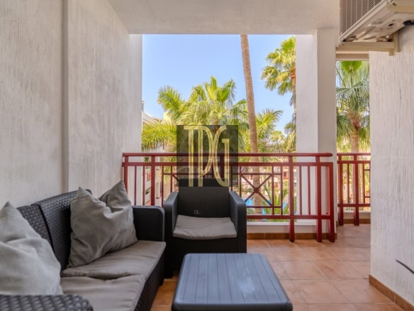 Balcony seating area with dark wicker sofa and armchair, beige cushions, a blue-gray coffee table, and a red railing with palm trees beyond.