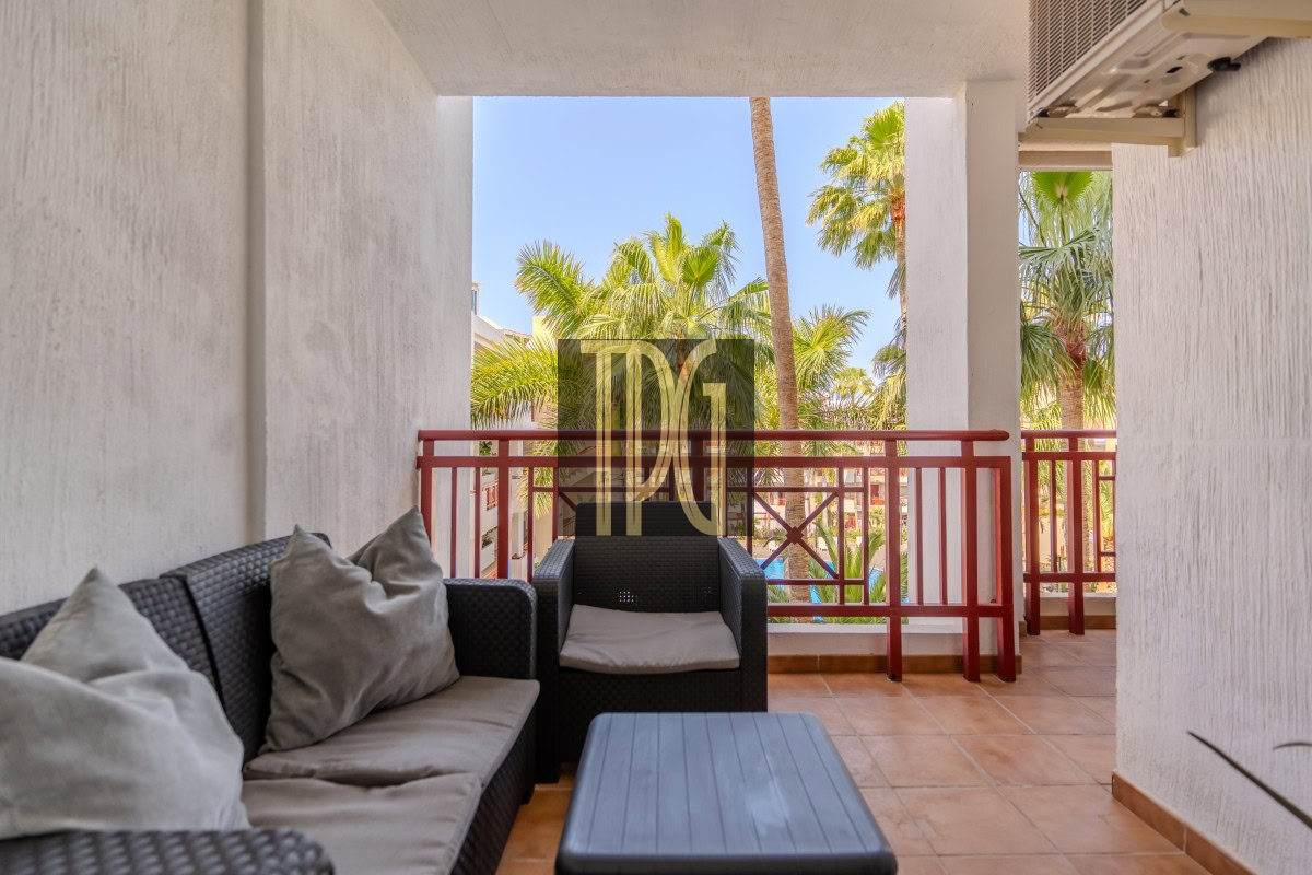 Balcony seating area with dark wicker sofa and armchair, beige cushions, a blue-gray coffee table, and a red railing with palm trees beyond.