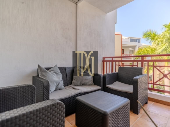 Outdoor balcony with dark wicker sofa set and light cushions, a matching chair and square coffee table, red railing and palm trees in the background.