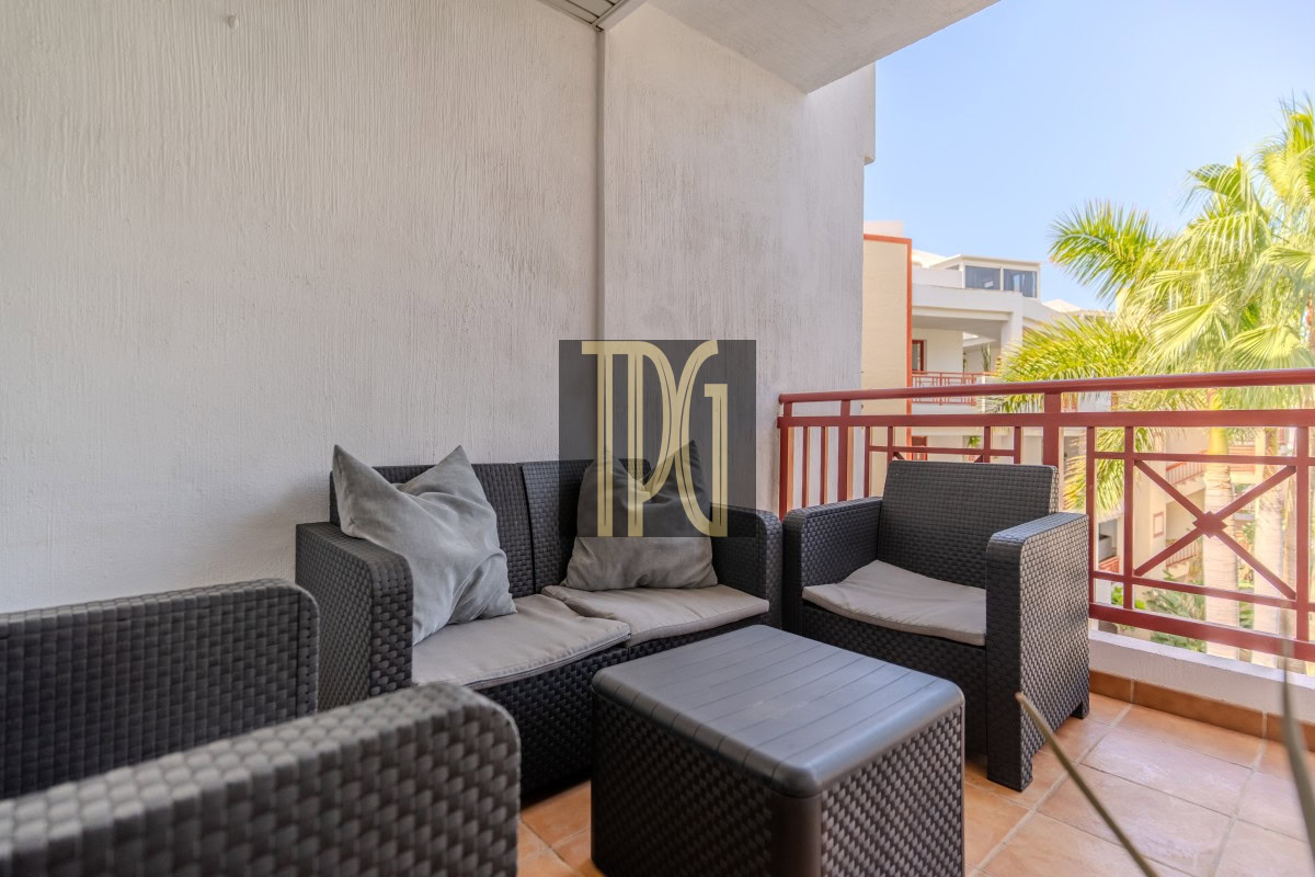 Outdoor balcony with dark wicker sofa set and light cushions, a matching chair and square coffee table, red railing and palm trees in the background.