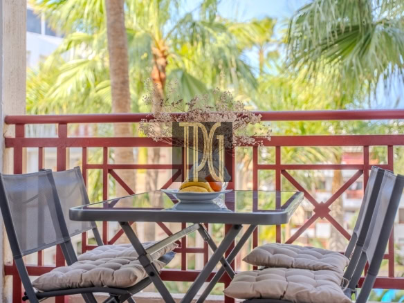 Balcony patio table with a bowl of fruit and four cushioned chairs, tropical palm trees in the background.