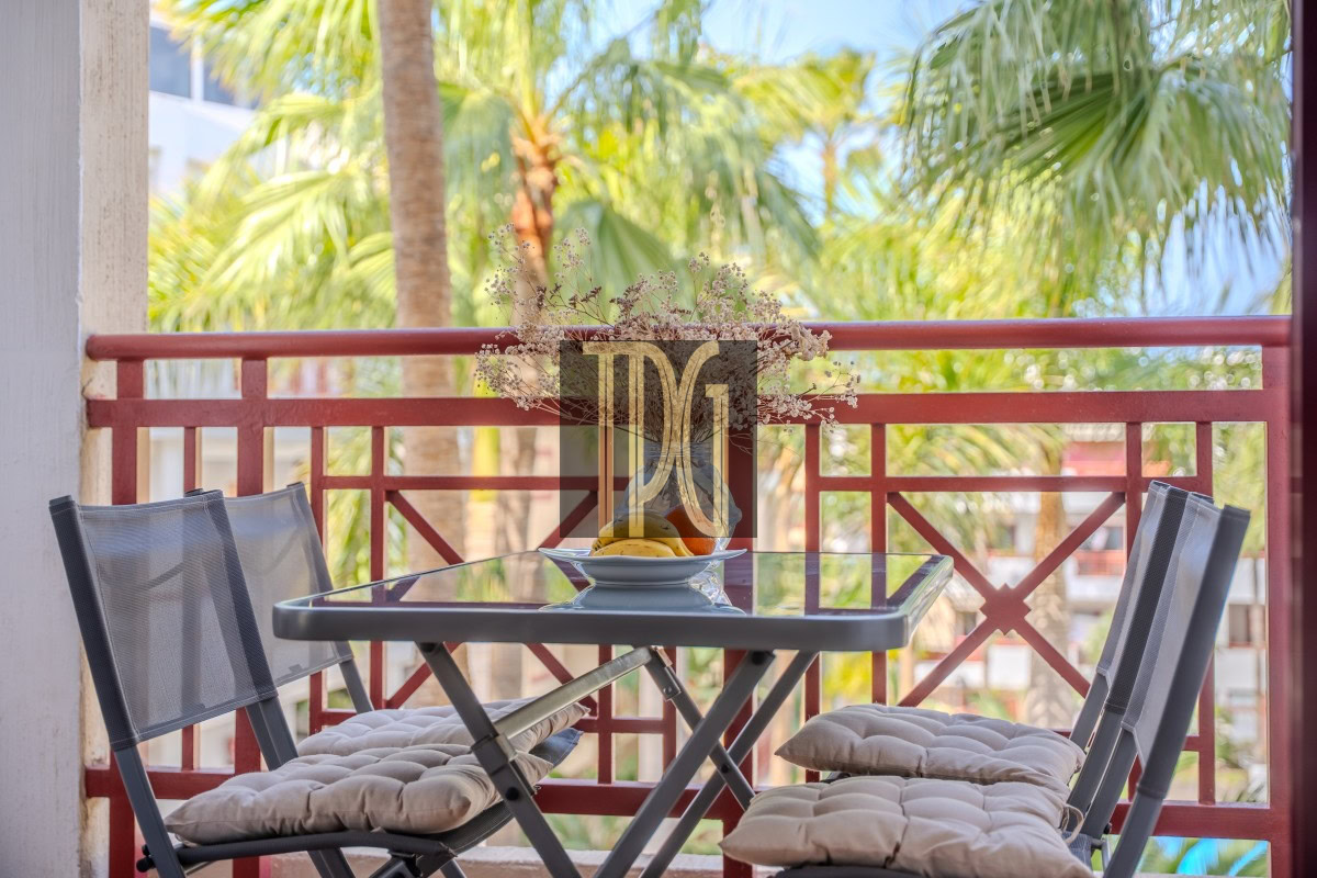 Balcony patio table with a bowl of fruit and four cushioned chairs, tropical palm trees in the background.
