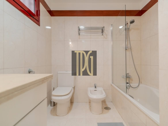 Modern bathroom with toilet, bidet, and bathtub/shower combo; beige tiled walls and a red-framed window above.