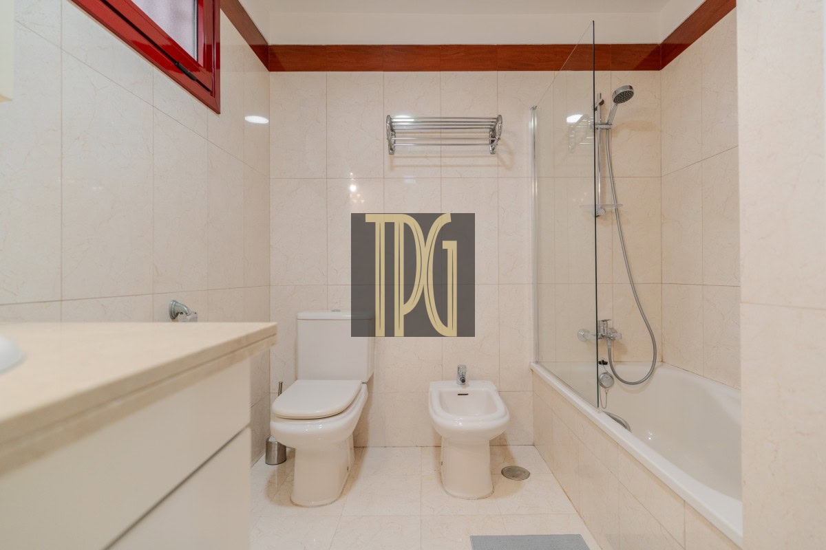 Modern bathroom with toilet, bidet, and bathtub/shower combo; beige tiled walls and a red-framed window above.
