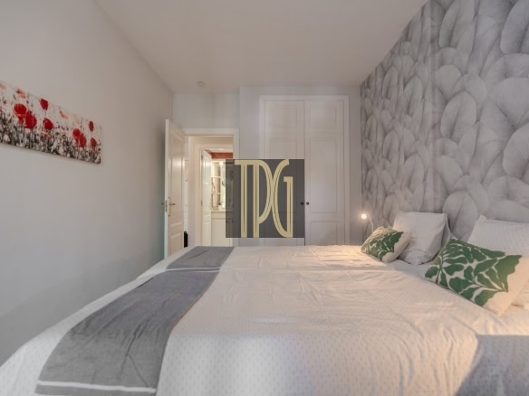 Hotel bedroom with a king-size bed, white linens, green leaf-pattern cushions, a grey textured wall, and a white wardrobe in the background.